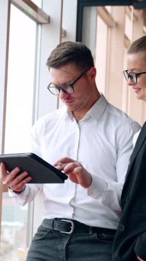 Ofiste çalışan meslektaşlar pencerede duruyorlar. Adam I-pad kullanıyor ve kadın aletin üzerindeki bir şeyi işaret ediyor. Dikey video