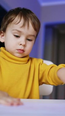 Güzel çocuk masa başında oturmuş resim çiziyor. Neşeli bebek mutlu bir şekilde gülümseyerek el kaldırır. Düşük açı. Kapatın. Dikey video