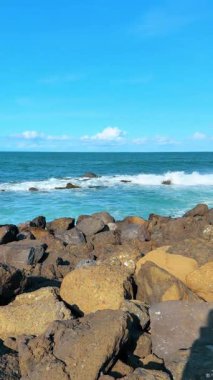 Büyük kayalar deniz kıyısını kaplar. Dalgalar kayalara çarpıyor ve köpüklü dalgalarla sıçrıyor. Dikey video.