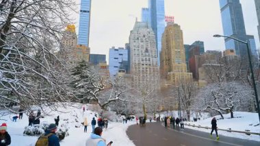 New York, ABD, 19 Aralık 2025: Birçok insan New York, ABD 'deki Central Park' ta asfalt yolda yürüyor ve koşuyor. Rickshaw 'lar karlı tepelerden yaklaşıyor. Manhattan mimarisi arkaplanda.