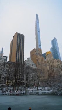 New York, ABD, 26 Aralık 2025 kışın Central Park, New York, ABD 'de kısmen buzla kaplı. Ağaçlar karla kaplı. Manhattan 'ın yüksek binalarının alçak açılı görüntüsü. Dikey video.