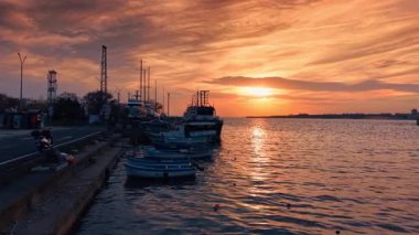 Güneş suyun üzerinde batarken tekneler kıyıya yakın bir marinada sıralanır. Gökyüzü turuncu ve pembe renkler gösteriyor, sakin yüzeyi yansıtıyor..