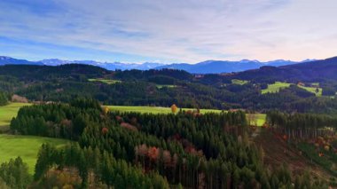 Geniş bir manzara bir dağ bölgesindeki yeşil tepeleri ve sık ormanları gösteriyor. Ağaçlarda sonbaharı gösteren turuncu ve sarı yapraklar var..