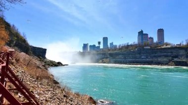 Ziyaretçiler Niagara Şelalesi yakınlarındaki patikalarda yürürken su kayalıklardan akıyor. Şehrin ufuk çizgisi arka planda parlak mavi bir gökyüzünün altında dimdik duruyor..
