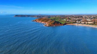 Geniş bir manzara karanın suyla buluştuğu bir kıyı şeridini gösteriyor. Sahilin arkasındaki tepeler yükselir ve kıyılar boyunca binalar görülebilir..