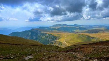 Dağlar bulutlu bir gökyüzünün altında yeşil tepeler boyunca uzanan bir patikayla yükselir. Yürüyüşçüler dar bir yolda, yeşil tepeler boyunca yürüyorlar. Dağlar uzaklarda ve bulutlar yukarıda..