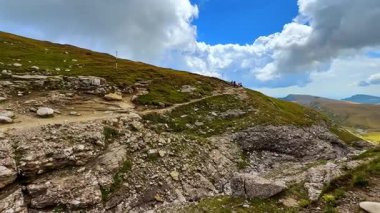 Yürüyüşçüler bulutların altındaki kayalık araziyi gün ışığında keşfederler. Bulutlu gökyüzüyle çevrili bir dağ yamacında iki kişi toprak patika boyunca yürür..