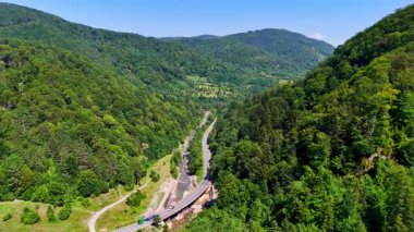 Açık mavi gökyüzünün altında yeşil tepelerden geçen yol. Yeşil tepeler ve ağaçlarla çevrili bir vadide nehir boyunca bir yol kıvrılır..