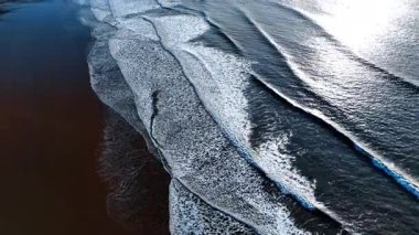 Koyu kahverengi kumlu bir kumsalla denizin ya da okyanusun kumlu sahillerinde uçmak. Geniş köpüklü dalgalar kıyıya doğru yavaşça akıyor..