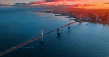 Birçok araba San Francisco-Oakland Bay Köprüsü 'nden geçiyor. Kırmızı ışık gün batımında şehir manzarasını kaplıyor. Birkaç gemi su manzarası üzerinde hareket ediyor. Hava görünümü.