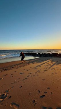 Kumda ayak izleriyle gün batımında kıyı şeridi boyunca yürümek. Güneş okyanusun üzerinde batarken bir insan sahilde yürür. Dalgalar kıyıya vurur ve ayak izleri kumu işaret eder..