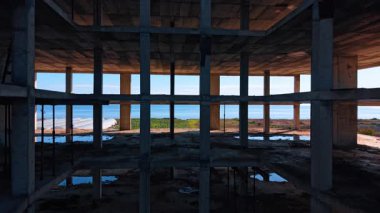 Tamamlanmamış binadaki boşluğun içine giriyoruz. Yapının zemininde bazı su birikintileri var. Arkaplanda deniz manzarası.