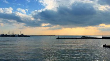 Gün batımında limanda tekneler görülürken bulutlar suyun üzerinde toplanır. Limana tekneler yanaşmış, bulutlar yukarıda ve gün batımında su ışığı yansıtıyor..