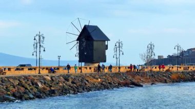 Burgaz, Bulgaristan, 29 Ekim 2025: İnsanlar sakin bir kasabada kıyıya yakın bir yel değirmeni kenarında yürüyorlar. Deniz kenarında açık bir günde insanlar bir yel değirmeninin yanındaki patikada gezinir.