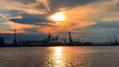 Yoğun bir limanın üzerinde gün batımında gemiler ve vinçler suyun üzerinde. Günbatımı bir limana ışık saçıyor, gökyüzünde vincler, gemiler ve bulutlar suya yansıyor..