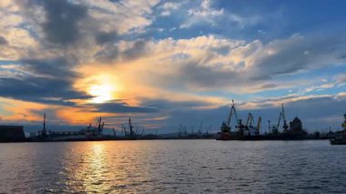 Limanda gün batımı vincler ve ışığı yansıtan suyla. Güneş su kenarındaki vinçlerin arkasında batarken gökyüzü renkleri sergiliyor. Tekneler sabit bulutlar üzerinde sürüklenirken.