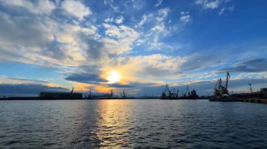 Uzakta tekneler ve vinçlerle dolu işlek bir limanın üzerinde akşam ışığı. Akşam saatlerinde bulutlar havada süzülürken, gün batımı limana ışık saçıyor. Tekneler ve turnalarla dolu..