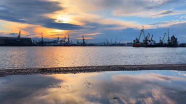 Limanda gün batımında su yansımaları ve vinçler görülebilir. Günbatımı renkleri limanın üstündeki gökyüzünü doldururken vinçler ve tekneler kıyı boyunca duruyor..