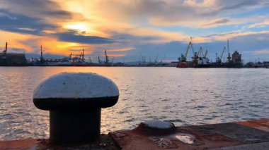 Limanın gün batımında demirleme bölmesi ve uzak tersane aktivitesi. Gemiler gün batımında uzakta çalışırken limanın kenarında bir demirleme kordonu duruyor..