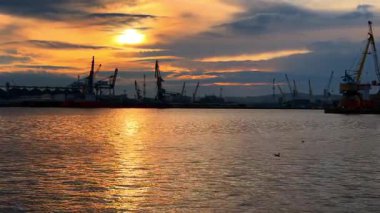 Endüstriyel limanda günbatımı, vinçlerle ve sakin su yansımalarıyla. Günbatımı gökyüzünü bir endüstri limanının üstündeki renklerle boyar..