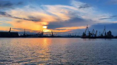 Suyun kenarında vinçlerle ve sanayi tekneleriyle dolu işlek bir limanda gün batımı. Gün batımında gökyüzü su yüzeyinde renk değiştirirken turnalar ve botlar bir limanda görülür..