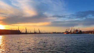 Gün batımında deniz kenarında gemiler ve turnalar. Vinçler ve gemiler limanda, güneş ufkun ardında renkli bulutlarla batarken görülür..