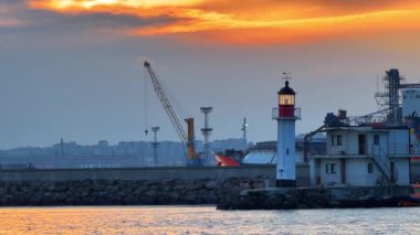 Limanda gün batımında deniz feneri ve uzak inşaat faaliyetleri var. Güneş batarken bir deniz feneri suyun kenarında duruyor..