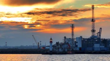 Arka planda deniz feneri ve sanayi yapıları olan liman üzerinde gün batımı. Güneş limanın arkasında görünür bir deniz feneri ve vinçle batıyor..