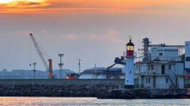Deniz feneri ve deniz kenarındaki binaların olduğu gün batımı manzarası. Sahne günbatımında bir limanı gösteriyor. Arka planda deniz feneri, binalar ve vinçler var..