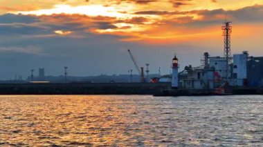 Deniz feneri ve sanayi manzaralı liman üzerinde gün batımı. Günbatımı, deniz feneri ve liman yapılarını aydınlatan turuncu ışığı suyun üzerine saçar..