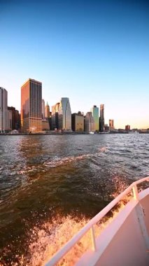 Dalgalar ve sıçramalar, su yüzeyinde hareket eden tekneden geliyor. New York gökdeleninde gün batımında. Dikey video.