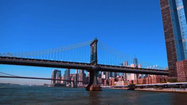 Manhattan Köprüsü 'nün ve Brooklyn Köprüsü' nün manzarası. Mavi açık gökyüzü ve Manhattan silueti. Nehir kıyısında iki tekne hareket ediyor..