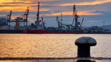 Sahil kenarında vincin ve gemilerin çalıştığı bir limanın günbatımı manzarası. Günbatımında işlek bir limana yanaşan bir gemi gibi turuncu ve pembe renkler gökyüzünü dolduruyor..