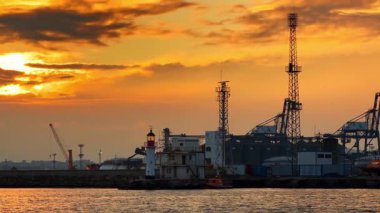 Vinçleri ve iletişim kuleleri olan bir liman manzarası. Sahne, gün batımında vinçler, iletişim kuleleri ve deniz feneri silueti olan bir limanı gösteriyor.