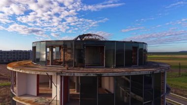 Silindir şeklindeki tamamlanmamış bina ile Koyu renk pencere panelleri. Arkaplanda mavi bulutlu gökyüzü ve su manzarası.