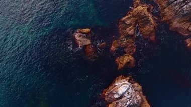 Rocky kıyı şeridi, gün ortasında kıyıda okyanus suyuyla buluşur. Dalgalar kıyıdaki kayalara çarparak parlak bir gökyüzünün altında mavi suyu ve doğal manzarayı gösterir..