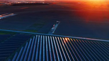 Güneş panelleri, güneş akşam ışığında manzaranın üzerinde batarken alanı kaplıyor. Güneş panelleri batan güneşin turuncu parıltısının altındaki alanı dolduruyor..