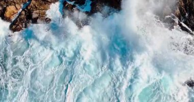 Kaya kıyısında köpüklü dalgalar hızla sıçrıyor. Su kayaların arasından akıyor. Üst görünüm.