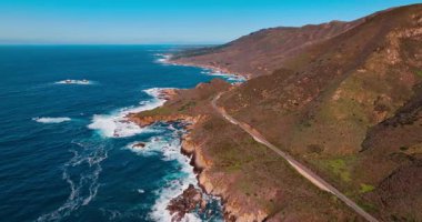 Çıplak dağların arasından geçen otoban. Güneşli bir günde, kayalık okyanus kıyısında İHA uçuşu. Big Sur, California, ABD.