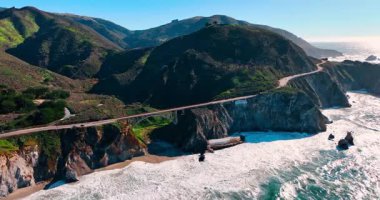 Köpüklü dalgalar sahile ve kumsala yuvarlanıyor. Arabalar, ABD 'nin Kaliforniya eyaletinin Big Sur kentindeki Bixby Köprüsü ve kayalıklar boyunca otoyoldan gidiyor. Hava görünümü.