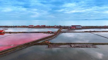Deniz kenarında tuz üretimi için çiftlikler. Dar kıyı şeridinde kırmızı çatılı binalar duruyor. Yoğun bulutlar gökyüzünü kaplıyor.