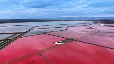 Kazıcı, büyük pembe tuz göletleriyle çevrili küçük bir toprak parçasında tuz topluyor. Tuz madenciliği için modern çiftlikler. Gri bulutlar gökyüzünü kaplıyor. Hava görünümü.