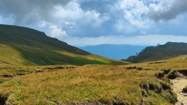 Gündüz vakti dağlarda bulutlu bir gökyüzünün altındaki çimenli tepelerin manzarası. Çimenli tepeler her iki taraftan yükseliyor. Gün boyunca bir dağ manzarasında bulutlar yükseliyor..