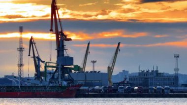 Limanda vinçleri ve kamyonları olan nakliye alanının günbatımı manzarası. Gün batımında rıhtımda vinçlerle bir gemi rıhtıma yanaştığı gibi kamyonlar diziliyor..