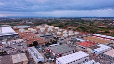 Petrol rafineri fabrikasına yaklaşan depoların üzerinden uçuş. Manzarada yakıt depolamak için büyük ve küçük tanklar var. Arkaplanda tarım alanları ve şehir manzarası.