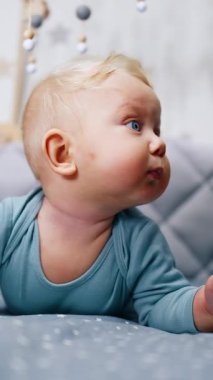 Beşiğinde küçük mavi gözlü, beyaz bir bebek var. Sevimli erkek bebek bir karyolanın tırabzanının üstünden bakmaya çalışıyor. Dikey video