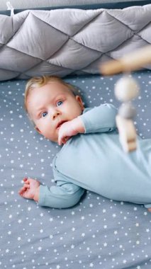 Mavi takım elbiseli sevimli bir bebek bebek beşikte yatar ve seyyar telefona bakar. Sevimli çocuk elini çiğneyip karyolada yuvarlanıyor. Üst Manzara. Dikey video