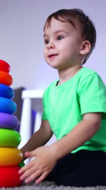 Koyu renk saçlı çocuk oyuncak piramidin önünde yerde oturuyor. Çocuk piramidi fırlatıp temelini atıyor. Dikey video