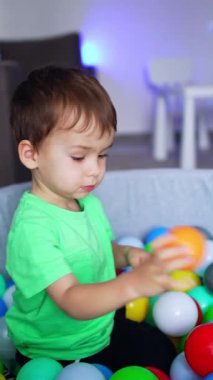 Tatlı çocuk elinde turuncu bir topla kuru havuzda oturuyor. Çocuk topu havuza atıyor ve havuzdan çıkıyor. Dikey video