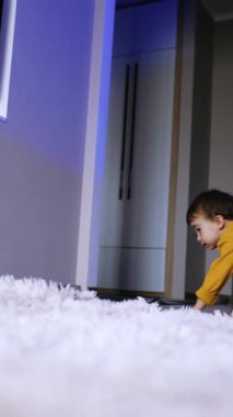 Sarı elbiseli enerjik beyaz çocuk neşeyle zıplıyor ve yerde yuvarlanıyor. Çocuk evde eğleniyor. Düşük açı. Dikey video
